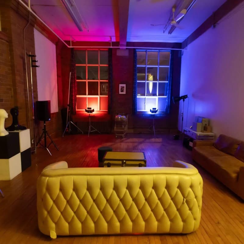 Content Day Studio at night: a loft with brick walls and wooden floors, two tall windows lit red and blue at the far end, a tufted sofa in front and a leather couch along the wall.