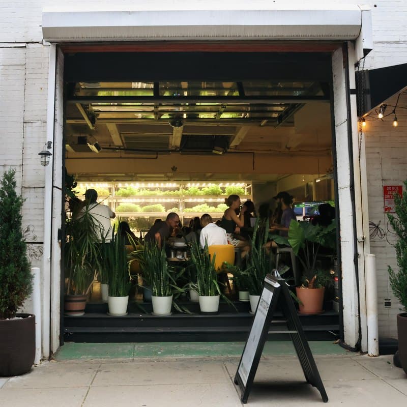 Farm.One from Bergen Street: the garage door rolled up on a room of people at tables, lit shelves of greens growing behind them, potted plants along the step.