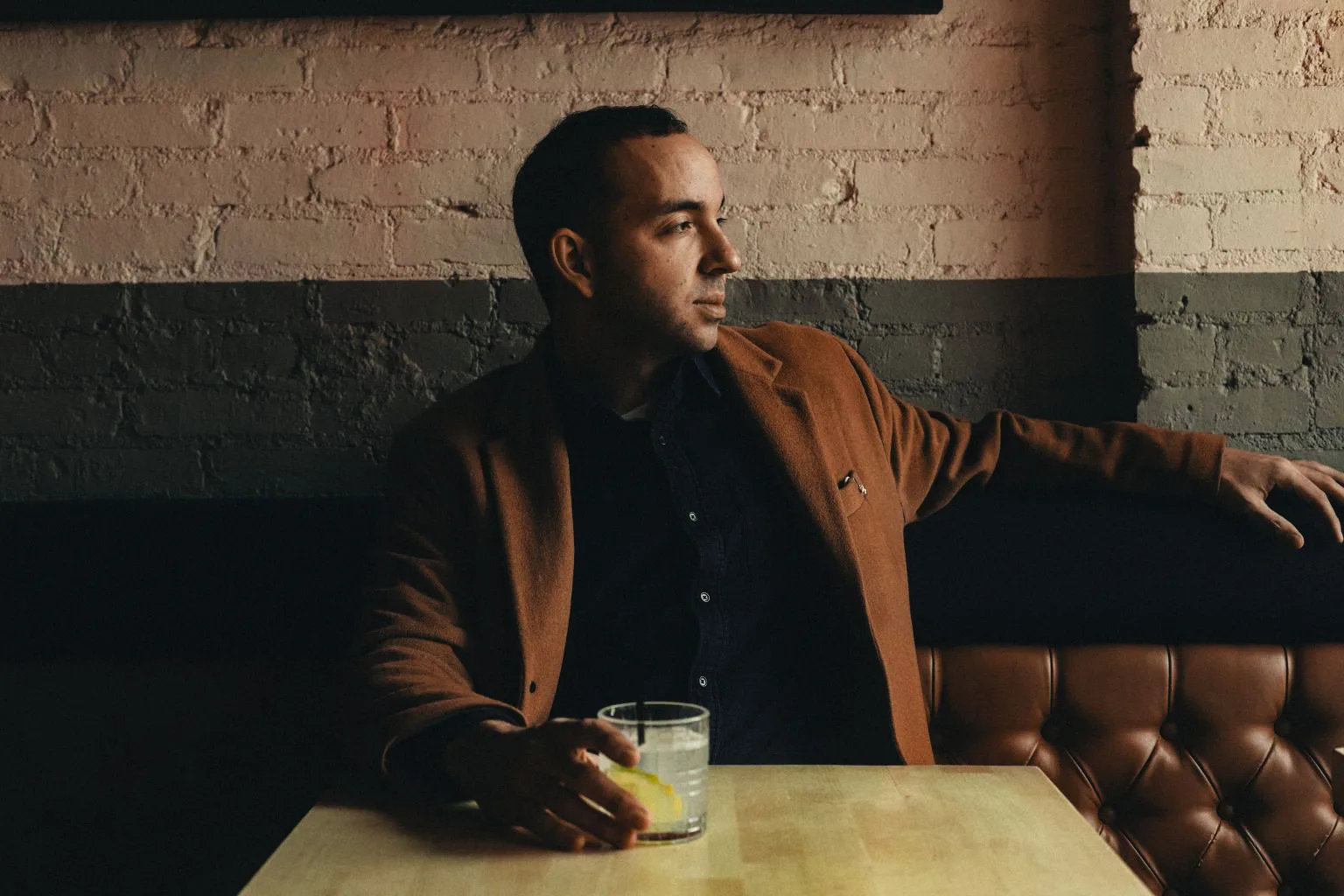 King Klavé seated in a tufted leather booth in a rust overshirt, one arm along the back, a drink with a lemon slice on the pale wood table, painted brick behind him