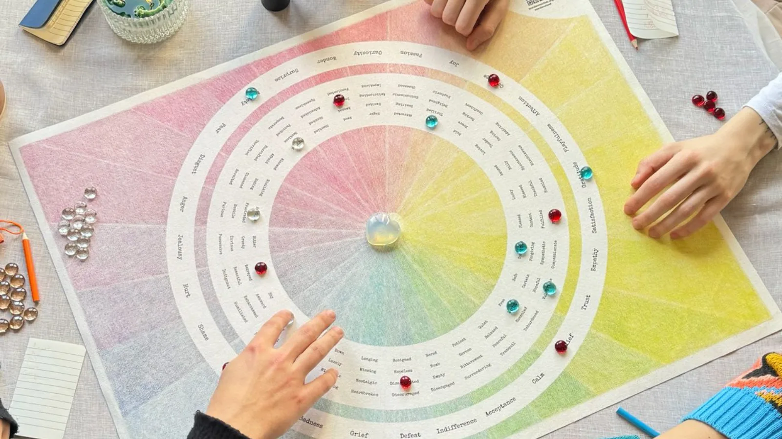 The WonderWheel board shot from above on pale linen, several pairs of hands reaching in, coloured glass markers set on emotion words around its rings and a glass heart at the centre