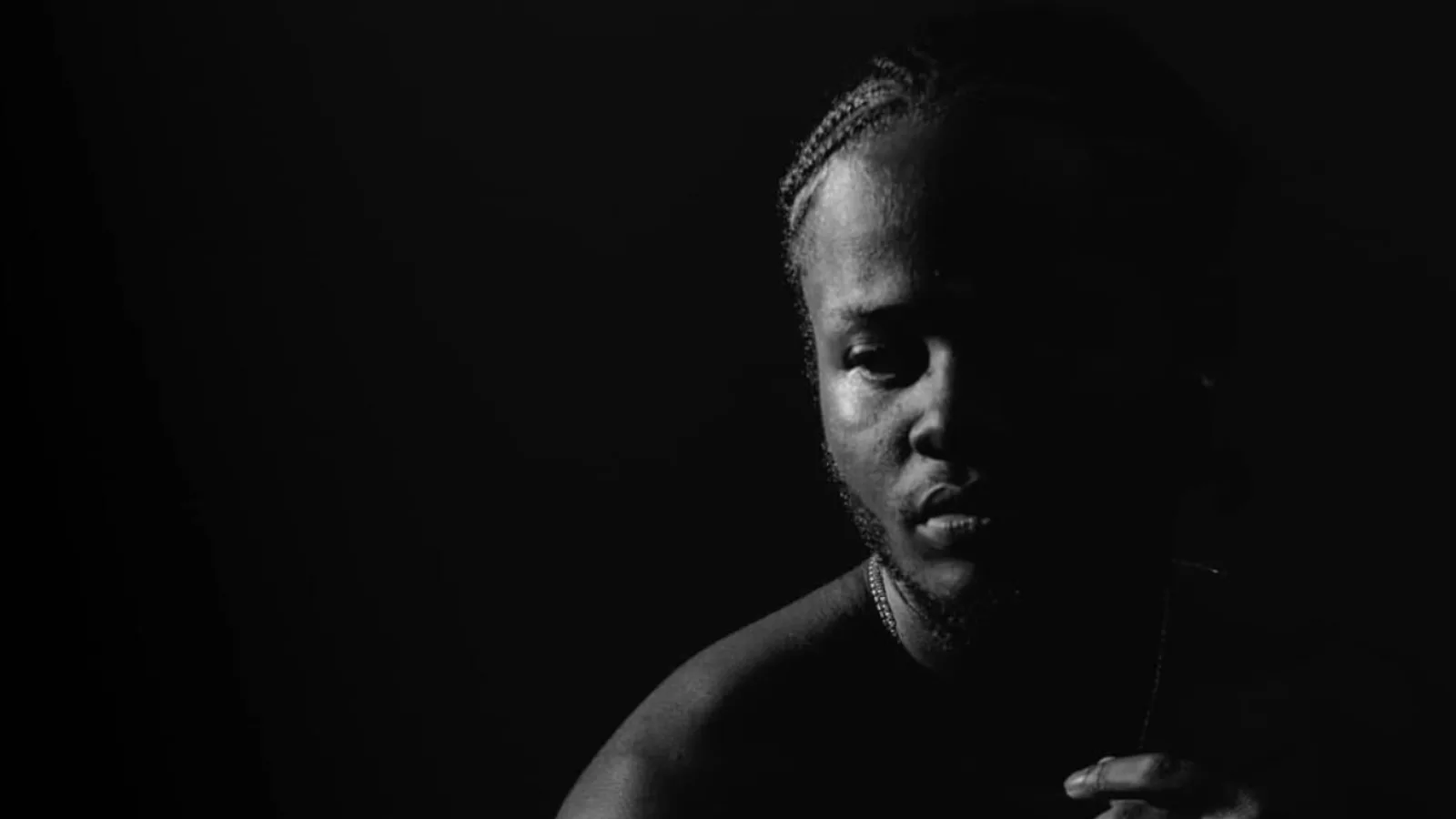 Ivan Cecil Walks in black and white, half his face caught in a rim of light against deep black, braids tied back and a chain at his collar